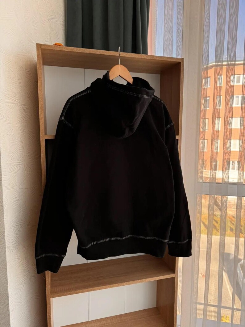 Pu&ograve; includere: Felpa con cappuccio nera con dettagli di cuciture bianche. La felpa &egrave; appesa a uno scaffale di legno.