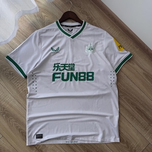 May include: White soccer jersey with green trim and accents. The jersey features the Newcastle United crest, sponsor logos, and the text "乐天堂 FUN88" in green. The jersey is hanging on a wooden hanger.