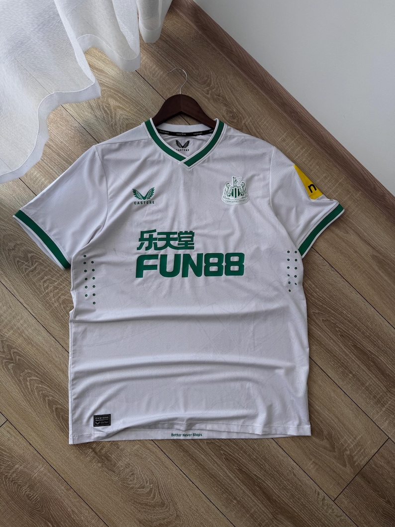 May include: White soccer jersey with green trim and accents. The jersey features the Newcastle United crest, sponsor logos, and the text "乐天堂 FUN88" in green. The jersey is hanging on a wooden hanger.