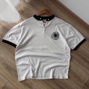 May include: A white short-sleeved shirt with black trim on the collar and sleeves. The shirt features a lace-up neckline with white laces and a circular black and white patch on the chest displaying an eagle emblem and the text Deutscher Fussball Bund.