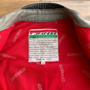 Op de afbeelding: Een rood leren jack met een zwarte kraag en een wit label met de tekst "Dainese" en een lijst met de namen van motorracekampioenen. Het label vermeldt ook "Made in Italy" en heeft het nummer 48.