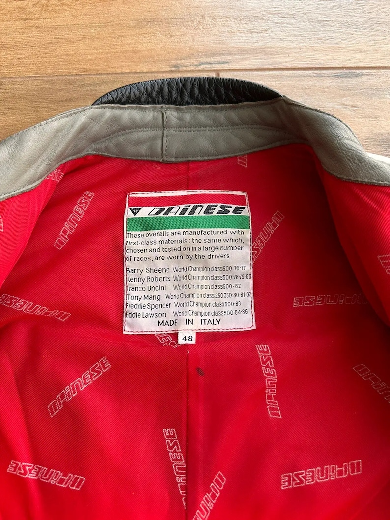 Op de afbeelding: Een rood leren jack met een zwarte kraag en een wit label met de tekst "Dainese" en een lijst met de namen van motorracekampioenen. Het label vermeldt ook "Made in Italy" en heeft het nummer 48.