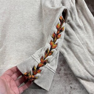 K&ouml;nnte beinhalten: Ein hellgraues Sweatshirt mit einem dekorativen, gestickten Blattdesign in Orange-, Gelb- und Braunt&ouml;nen am &Auml;rmel. Das Sweatshirt hat einen gerippten Bund und eine lockere Passform.