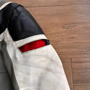 Op de afbeelding: Een grijs, wit en zwart leren jack met een rode accent op de mouw. Het jack heeft een ritssluiting en een opstaande kraag.