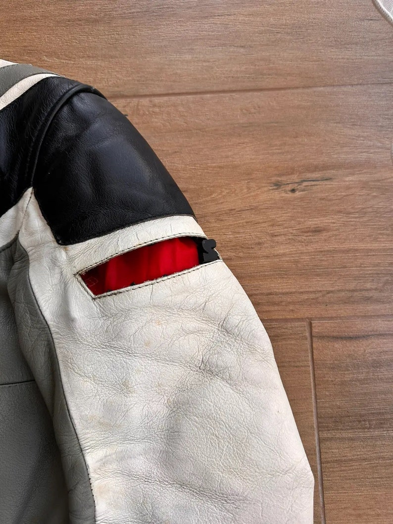 Op de afbeelding: Een grijs, wit en zwart leren jack met een rode accent op de mouw. Het jack heeft een ritssluiting en een opstaande kraag.