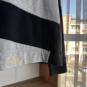 Op de afbeelding: Een zwart-grijs gestreept sweatshirt met een geborduurd logo in goud met de tekst "Danielle 10". Het sweatshirt hangt aan een houten plank.