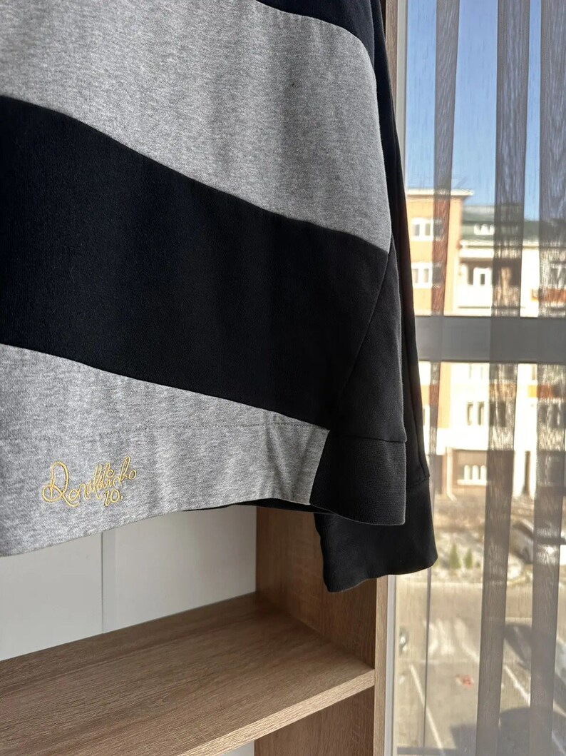 Op de afbeelding: Een zwart-grijs gestreept sweatshirt met een geborduurd logo in goud met de tekst "Danielle 10". Het sweatshirt hangt aan een houten plank.