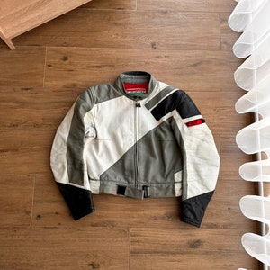 Op de afbeelding: Een wit, grijs en zwart leren motorjack met een ritssluiting en een riem in de taille. Het jack heeft een geometrisch patroon en het woord "Dainese" staat gedrukt op de linker borst.