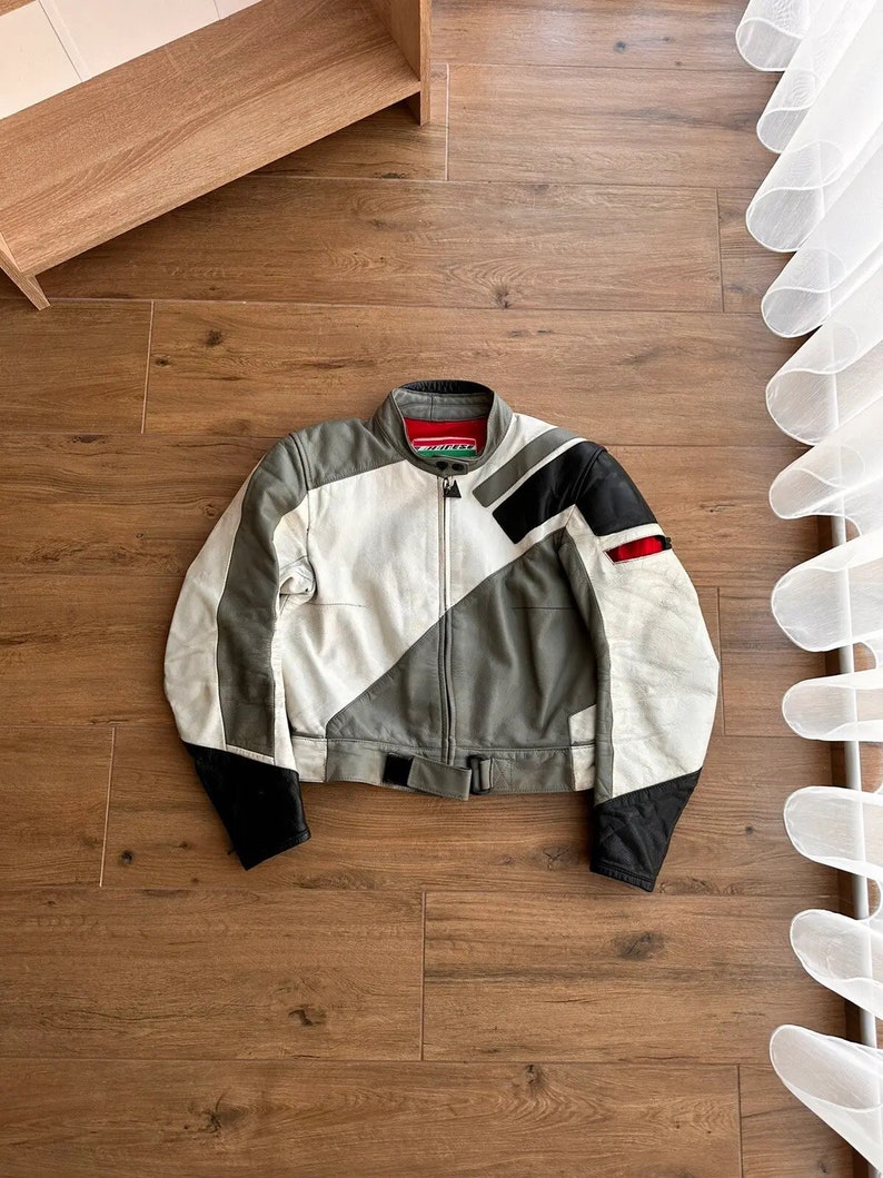 Op de afbeelding: Een wit, grijs en zwart leren motorjack met een ritssluiting en een riem in de taille. Het jack heeft een geometrisch patroon en het woord "Dainese" staat gedrukt op de linker borst.