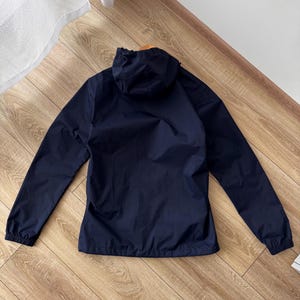 May include: A navy blue hooded jacket with long sleeves. The jacket is made of a lightweight material and has a drawstring hood. The cuffs and hem are elasticated. The jacket is on a wooden floor.