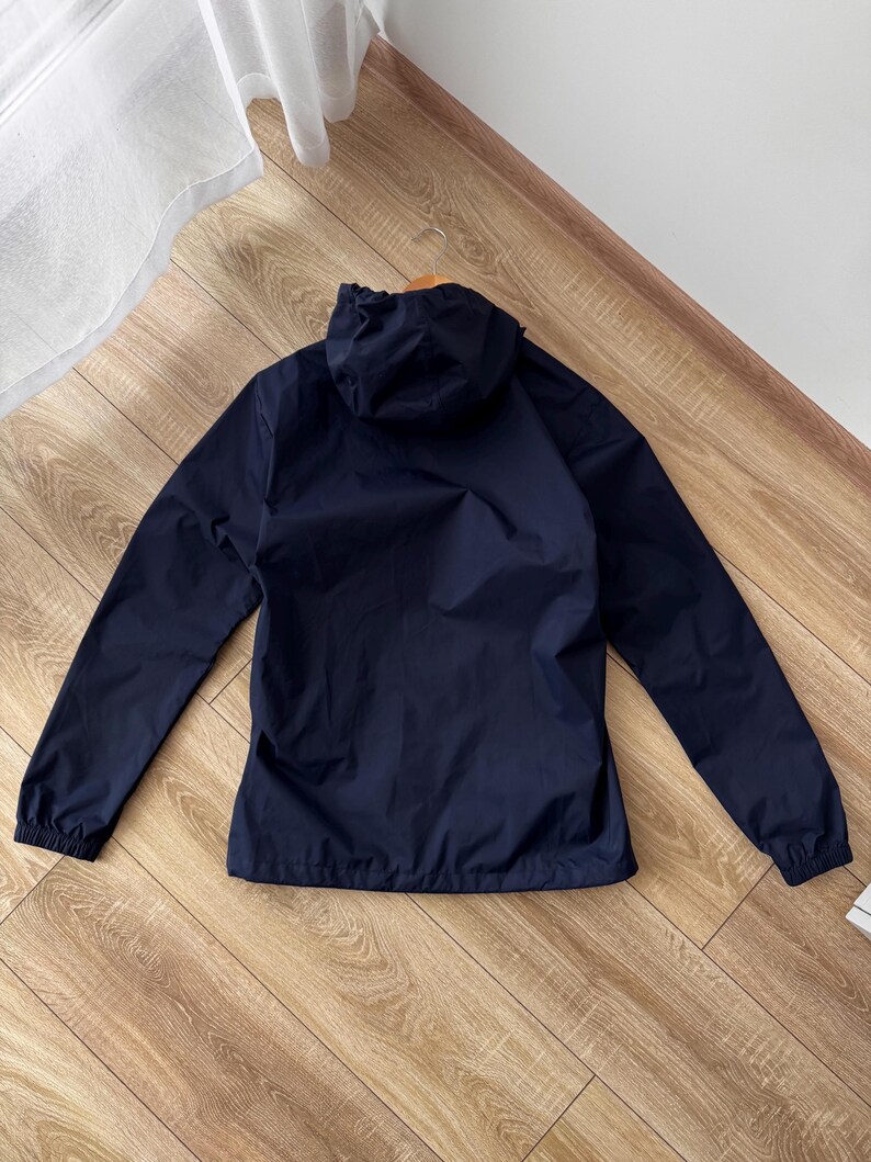 May include: A navy blue hooded jacket with long sleeves. The jacket is made of a lightweight material and has a drawstring hood. The cuffs and hem are elasticated. The jacket is on a wooden floor.