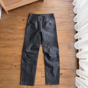 Pu&ograve; includere: Pantaloni in pelle nera con design a gamba dritta. I pantaloni hanno chiusura con bottone e cerniera, passanti per cintura e tasche. La pelle appare leggermente strutturata. I pantaloni sono appoggiati piatti su una superficie di legno.