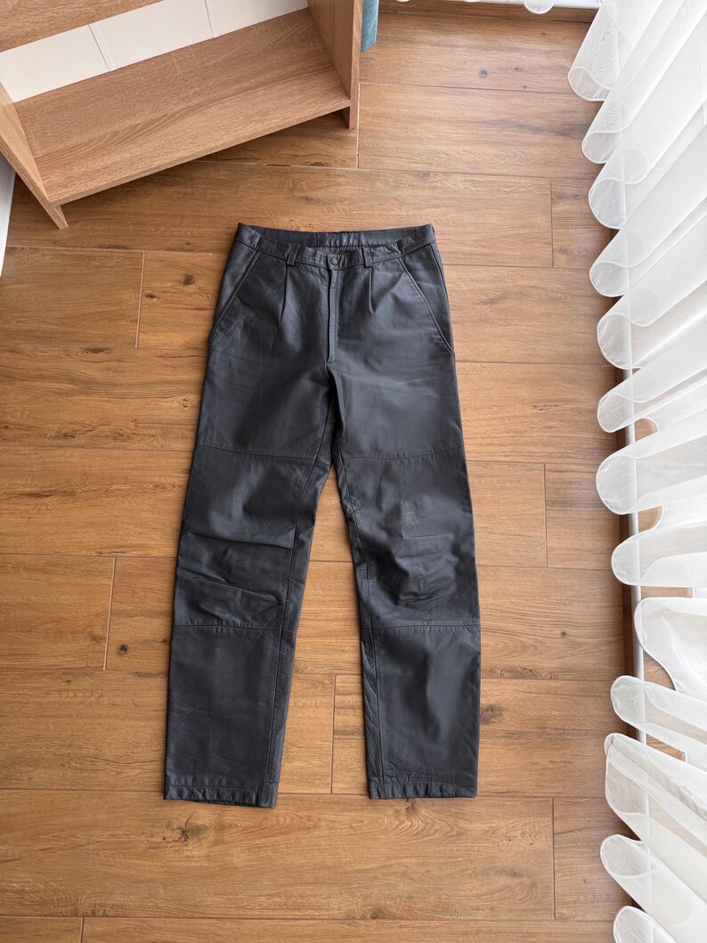 Pu&ograve; includere: Pantaloni in pelle nera con design a gamba dritta. I pantaloni hanno chiusura con bottone e cerniera, passanti per cintura e tasche. La pelle appare leggermente strutturata. I pantaloni sono appoggiati piatti su una superficie di legno.