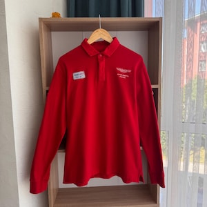 Puede incluir: Un polo rojo brillante de manga larga con cuello y cierre de botones. La camisa presenta dos parches: uno con el texto "SBB" y otro con "ASTON MARTIN RACING" y un logotipo. La camisa está colgada en una percha de madera.