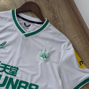 May include: White soccer jersey with green trim and the Newcastle United crest. The jersey has the Castore logo on the chest and sleeve. The jersey also has the text "FUN88" in green.