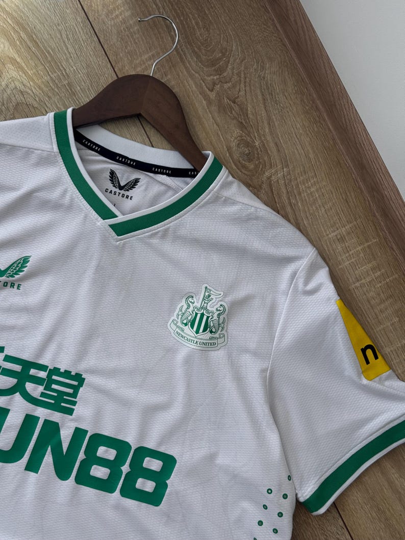 May include: White soccer jersey with green trim and the Newcastle United crest. The jersey has the Castore logo on the chest and sleeve. The jersey also has the text "FUN88" in green.