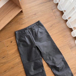 Pu&ograve; includere: Pantaloni in pelle nera dal design classico. I pantaloni hanno chiusura con bottone e zip, passanti per cintura e tasche. La pelle sembra morbida ed elastica. I pantaloni sono appoggiati su un pavimento in legno.