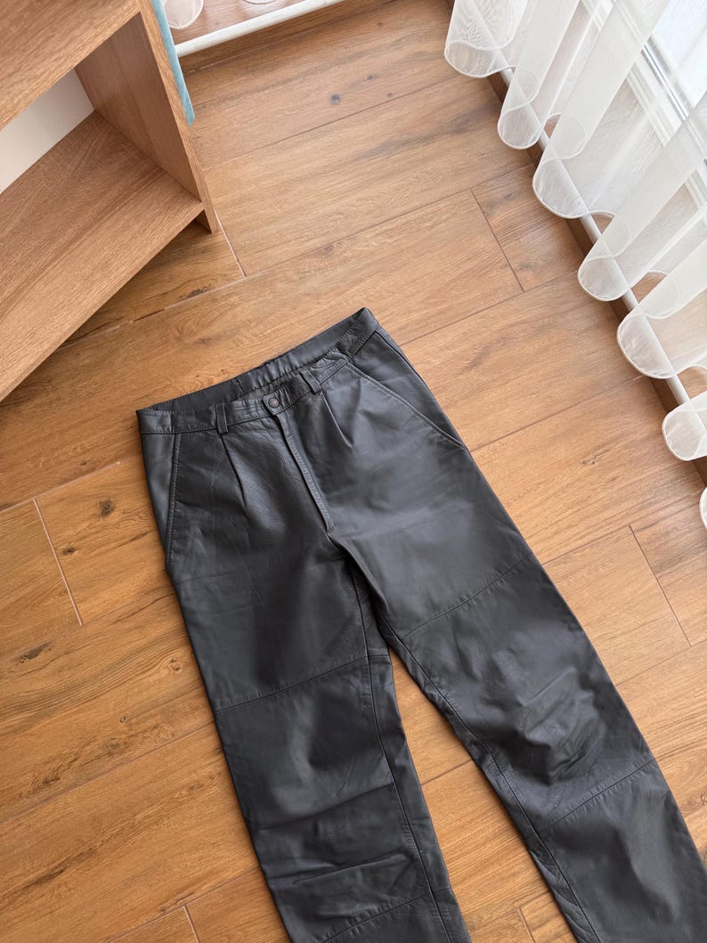 Pu&ograve; includere: Pantaloni in pelle nera dal design classico. I pantaloni hanno chiusura con bottone e zip, passanti per cintura e tasche. La pelle sembra morbida ed elastica. I pantaloni sono appoggiati su un pavimento in legno.