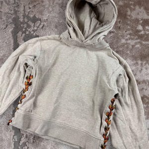 K&ouml;nnte beinhalten: Ein hellgrauer Kapuzenpullover mit Kordelzug und langen &Auml;rmeln. Die Seiten des Hoodies weisen ein dekoratives, farbenfrohes, gewebtes Design in Orange-, Gelb- und Braunt&ouml;nen auf. Der Hoodie ist aus einem weichen, bequemen Material gefertigt.