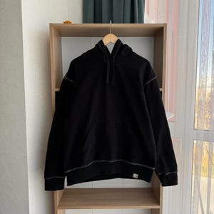 Pu&ograve; includere: Felpa con cappuccio nera con una tasca frontale e una piccola patch con logo in basso a destra. La felpa ha un cappuccio con coulisse ed &egrave; appesa a uno scaffale in legno.