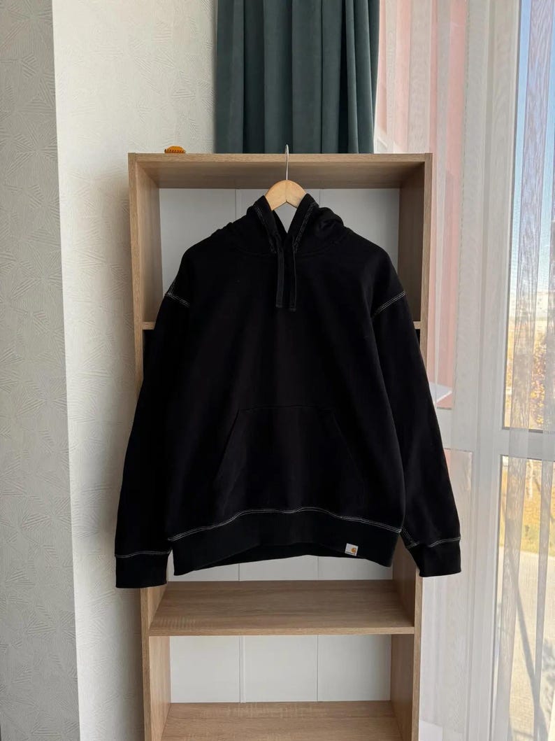 Pu&ograve; includere: Felpa con cappuccio nera con una tasca frontale e una piccola patch con logo in basso a destra. La felpa ha un cappuccio con coulisse ed &egrave; appesa a uno scaffale in legno.
