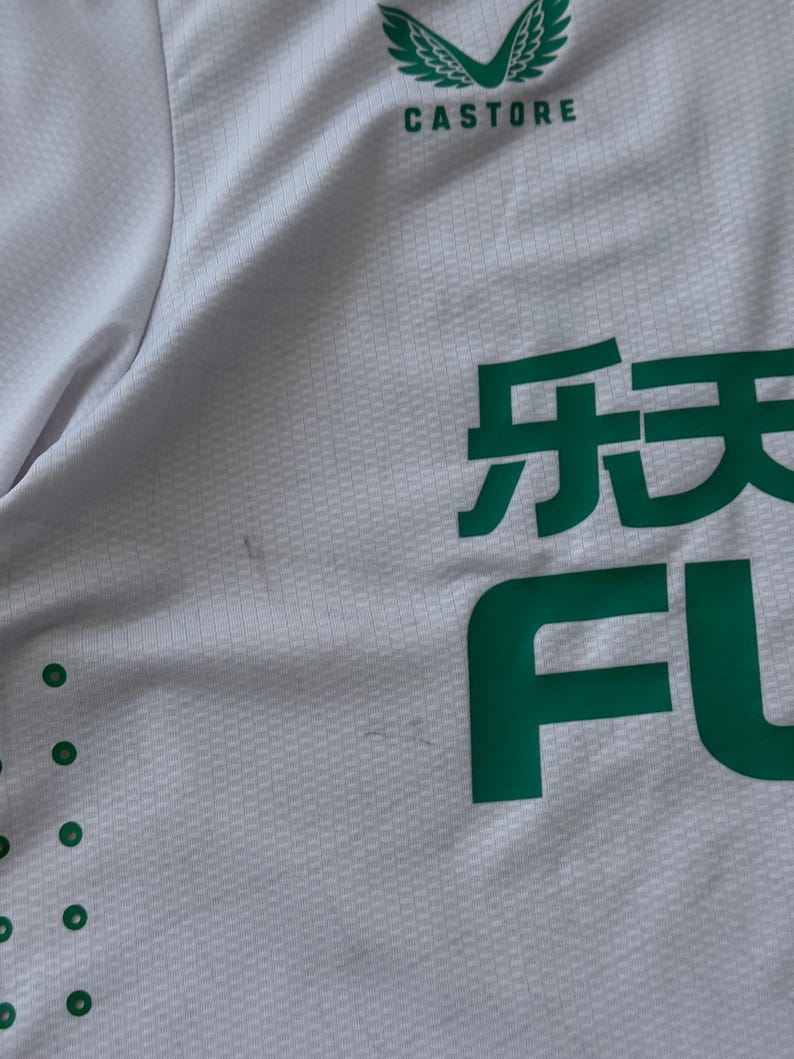 May include: White athletic jersey with green accents. The jersey features the brand name "Castore" in green at the top and large green Japanese characters and the letters "FU" on the front. The fabric has a subtle textured pattern.