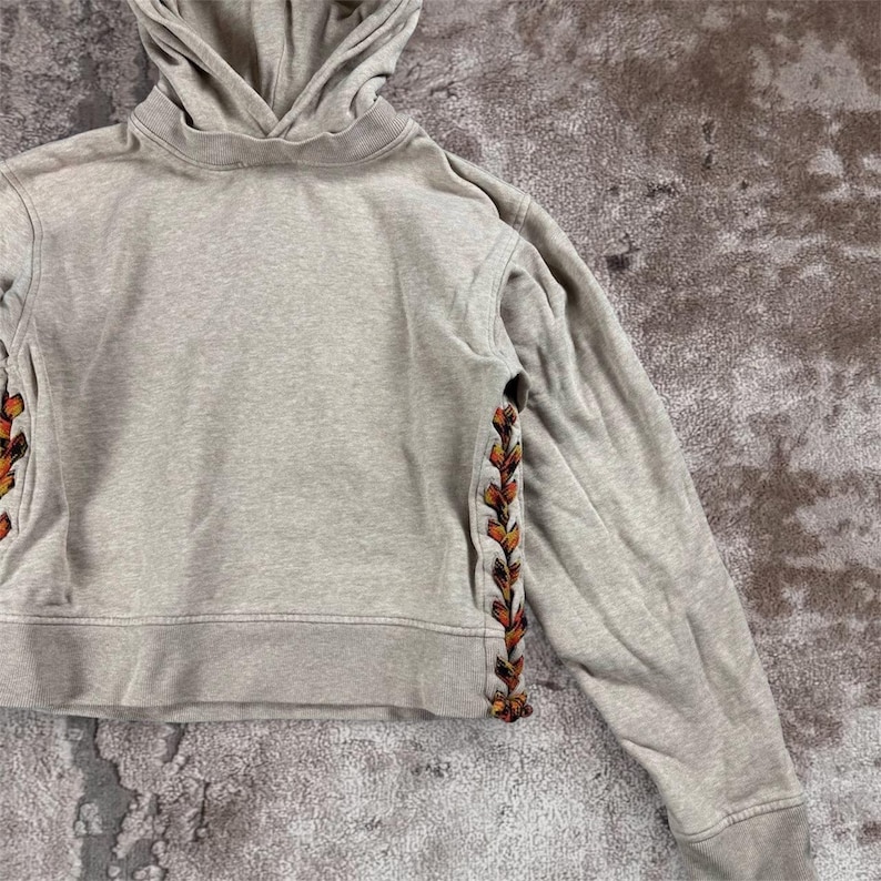 K&ouml;nnte beinhalten: Ein hellgrauer Kapuzenpullover mit Kordelzugkapuze und langen &Auml;rmeln. Die Seiten des Hoodies weisen ein dekoratives, farbenfrohes, gesticktes Design auf. Der Hoodie ist aus einem weichen, bequemen Material gefertigt.
