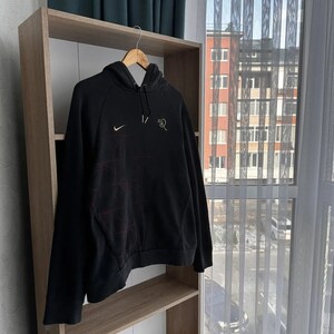 Op de afbeelding: Zwarte hoodie met een wit Nike swoosh logo en rode stiksels aan de linkerkant van de borst. De tekst "1/2" is in wit gedrukt aan de rechterkant van de borst.