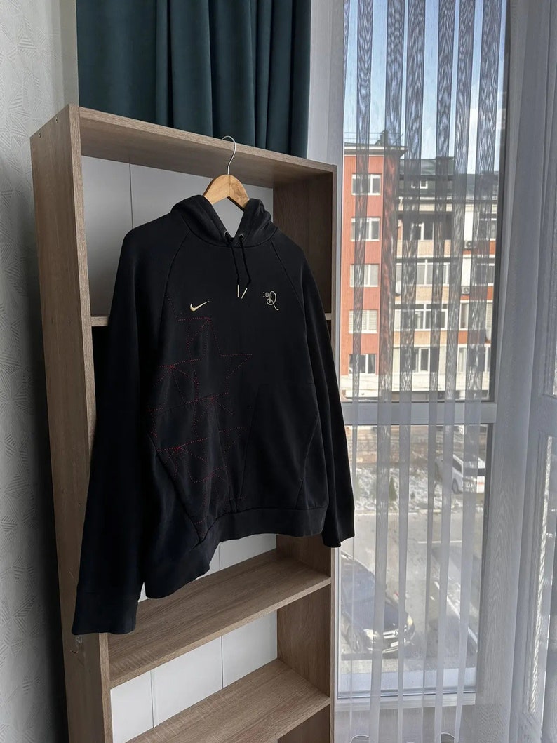 Op de afbeelding: Zwarte hoodie met een wit Nike swoosh logo en rode stiksels aan de linkerkant van de borst. De tekst "1/2" is in wit gedrukt aan de rechterkant van de borst.