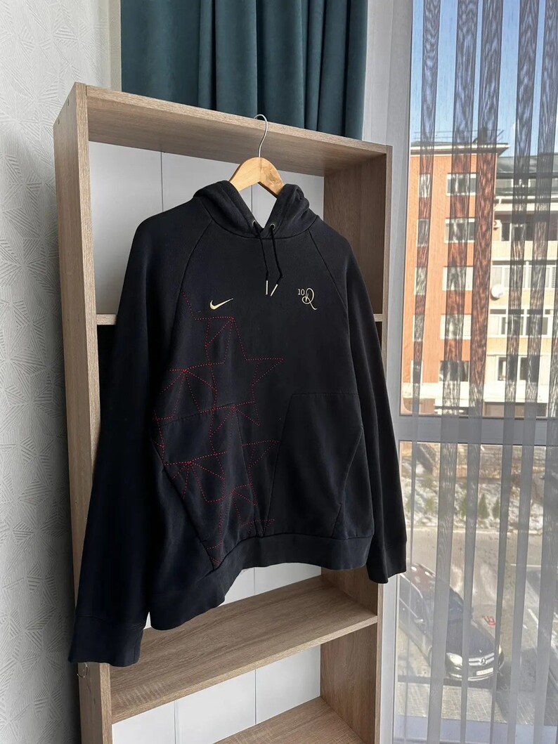 Op de afbeelding: Zwarte hoodie met een rood en wit sterrenontwerp en een Nike-logo op de linkerborst. De tekst "1/2" staat op de rechterborst.