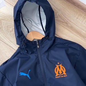May include: Navy blue hooded jacket with a full zip. The jacket displays a blue Puma logo and an orange emblem with the text "DROIT AU BUT". The hood has a drawstring and a light-coloured lining. A wooden hanger is visible inside the hood.