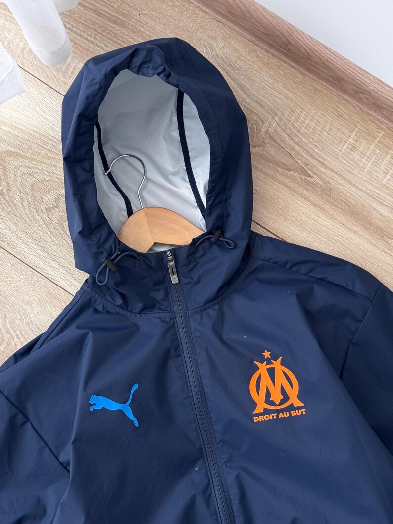 May include: Navy blue hooded jacket with a full zip. The jacket displays a blue Puma logo and an orange emblem with the text "DROIT AU BUT". The hood has a drawstring and a light-coloured lining. A wooden hanger is visible inside the hood.
