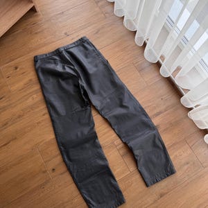 Pu&ograve; includere: Pantaloni in pelle nera dal design classico. I pantaloni hanno un taglio dritto e presentano dettagli di cucitura visibili. L'indumento &egrave; appoggiato su un pavimento in legno, mostrando la sua consistenza e forma.