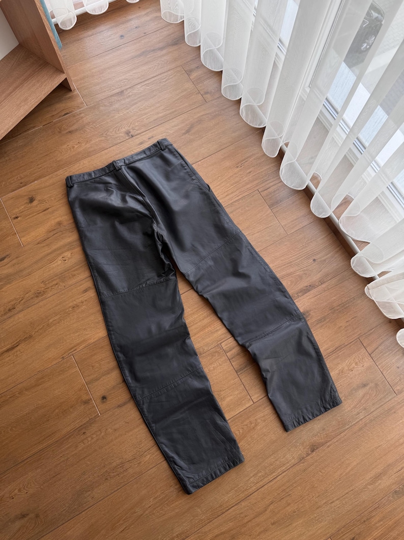 Pu&ograve; includere: Pantaloni in pelle nera dal design classico. I pantaloni hanno un taglio dritto e presentano dettagli di cucitura visibili. L'indumento &egrave; appoggiato su un pavimento in legno, mostrando la sua consistenza e forma.