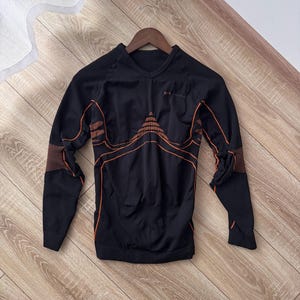 Puede incluir: Camiseta deportiva negra de manga larga con líneas y detalles en naranja. La camiseta tiene cuello en V y un logotipo en el pecho. Las mangas tienen una textura acanalada en los puños. La camiseta está colgada de una percha de madera.