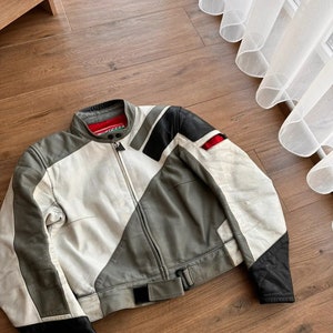 Op de afbeelding: Een wit, grijs en zwart leren motorjack met een ritssluiting en een riem. Het jack heeft een rood label op de kraag.