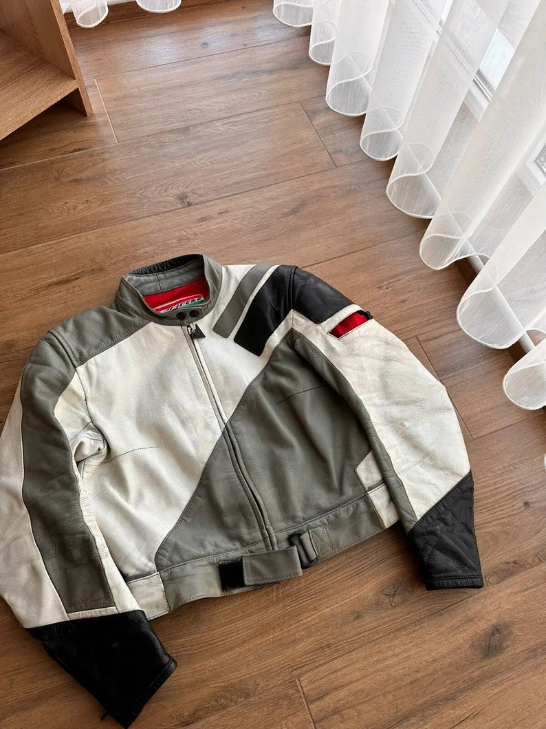 Op de afbeelding: Een wit, grijs en zwart leren motorjack met een ritssluiting en een riem. Het jack heeft een rood label op de kraag.