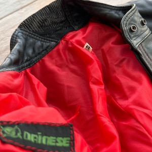 May include: Black leather jacket with a red lining and a green label that says "Driinese". The jacket has a zipper closure and a ribbed collar.