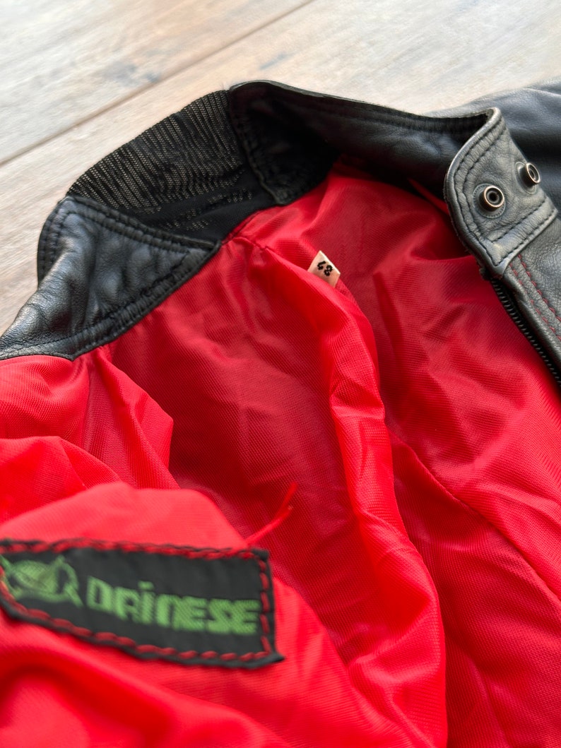 May include: Black leather jacket with a red lining and a green label that says "Driinese". The jacket has a zipper closure and a ribbed collar.