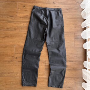 Pu&ograve; includere: Pantaloni grigio scuro con design a gamba dritta. I pantaloni sembrano essere realizzati in un materiale simile alla pelle e presentano cuciture visibili. I pantaloni sono appoggiati su una superficie di legno.