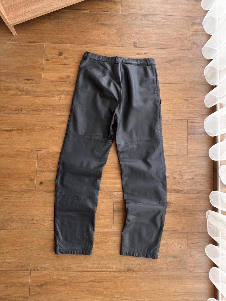 Pu&ograve; includere: Pantaloni grigio scuro con design a gamba dritta. I pantaloni sembrano essere realizzati in un materiale simile alla pelle e presentano cuciture visibili. I pantaloni sono appoggiati su una superficie di legno.