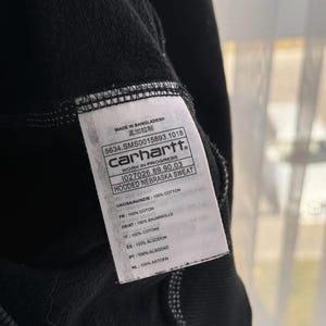 Pu&ograve; includere: Un'etichetta di felpa con cappuccio Carhartt nera con testo bianco. L'etichetta riporta "Made in Bangladesh", "5634.SMS0015893.1018", "Carhartt", "Work in Progress", "1027026.89.90.03", "Hooded Nebraska Sweat", e elenca il contenuto del tessuto in pi&ugrave; lingue.