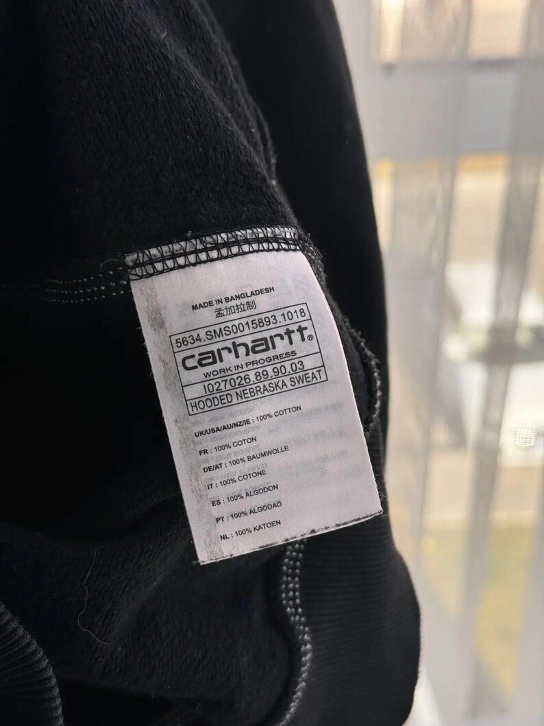 Pu&ograve; includere: Un'etichetta di felpa con cappuccio Carhartt nera con testo bianco. L'etichetta riporta "Made in Bangladesh", "5634.SMS0015893.1018", "Carhartt", "Work in Progress", "1027026.89.90.03", "Hooded Nebraska Sweat", e elenca il contenuto del tessuto in pi&ugrave; lingue.