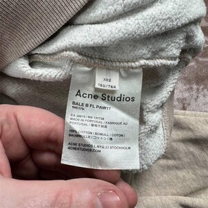 K&ouml;nnte beinhalten: Nahaufnahme eines Kleidungsst&uuml;cketiketts von Acne Studios. Das Etikett ist wei&szlig; mit schwarzem Text, einschlie&szlig;lich des Markennamens, der Gr&ouml;&szlig;e XXS und der Stoffzusammensetzung. Das Kleidungsst&uuml;ck ist hellbeige.