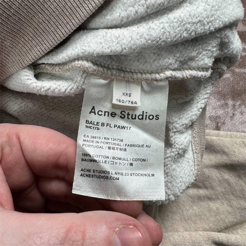 K&ouml;nnte beinhalten: Nahaufnahme eines Kleidungsst&uuml;cketiketts von Acne Studios. Das Etikett ist wei&szlig; mit schwarzem Text, einschlie&szlig;lich des Markennamens, der Gr&ouml;&szlig;e XXS und der Stoffzusammensetzung. Das Kleidungsst&uuml;ck ist hellbeige.