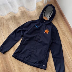 May include: Navy blue hooded jacket with a full-length zip. The jacket features a blue Puma logo and an orange and blue emblem on the chest. The jacket is made of a lightweight material and has a hood.