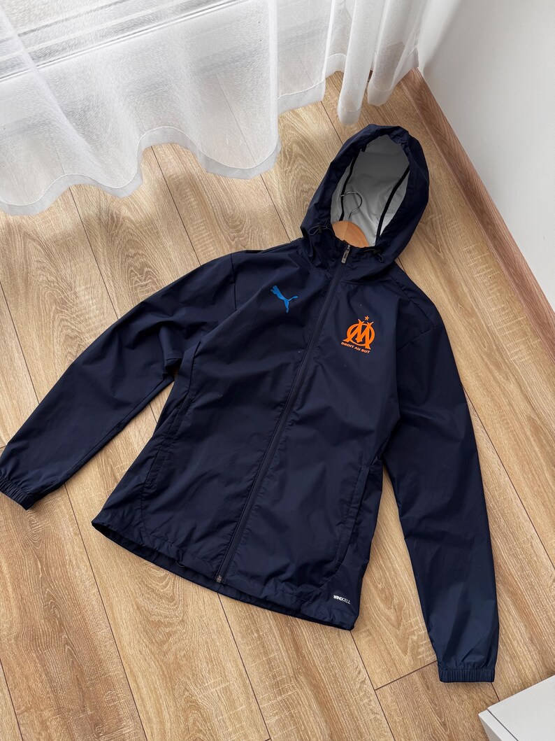 May include: Navy blue hooded jacket with a full-length zip. The jacket features a blue Puma logo and an orange and blue emblem on the chest. The jacket is made of a lightweight material and has a hood.