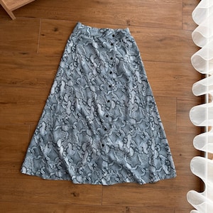 Puede incluir: Una falda larga de color azul con estampado de serpiente y cierre de botones en la parte delantera.