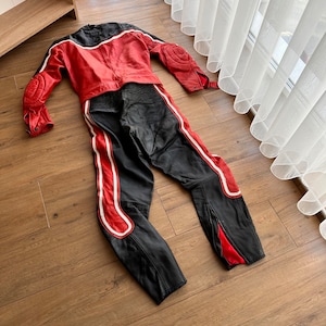 May include: A red and black leather motorcycle racing suit with white accents. The suit is laid out flat on a wooden floor.