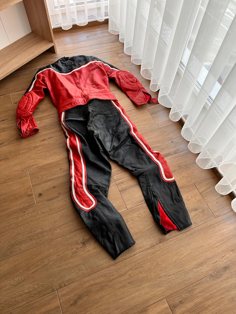May include: A red and black leather motorcycle racing suit with white accents. The suit is laid out flat on a wooden floor.